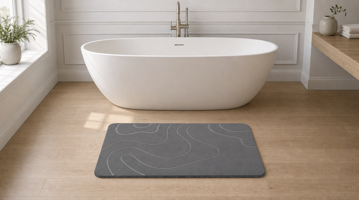 Gray stone bath mat with broad abstract curved lines on a light wood-look floor in front of a white tub.
