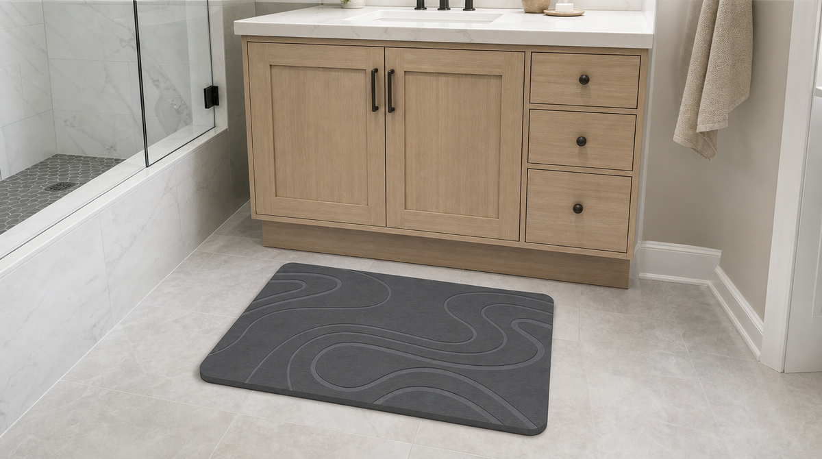Dark gray stone bath mat with flowing nested curves in front of a wood vanity beside a glass shower.