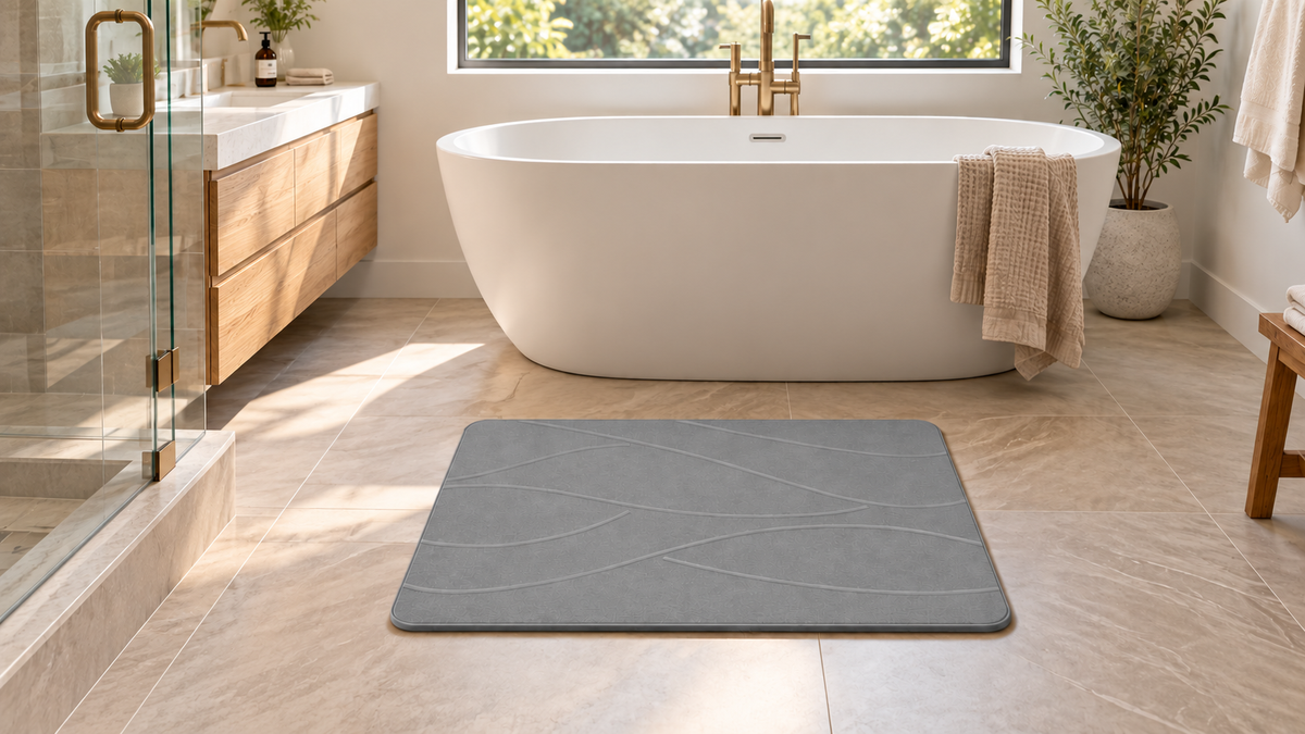Gray stone bath mat with fine curved lines on beige tile in front of a freestanding bathtub.