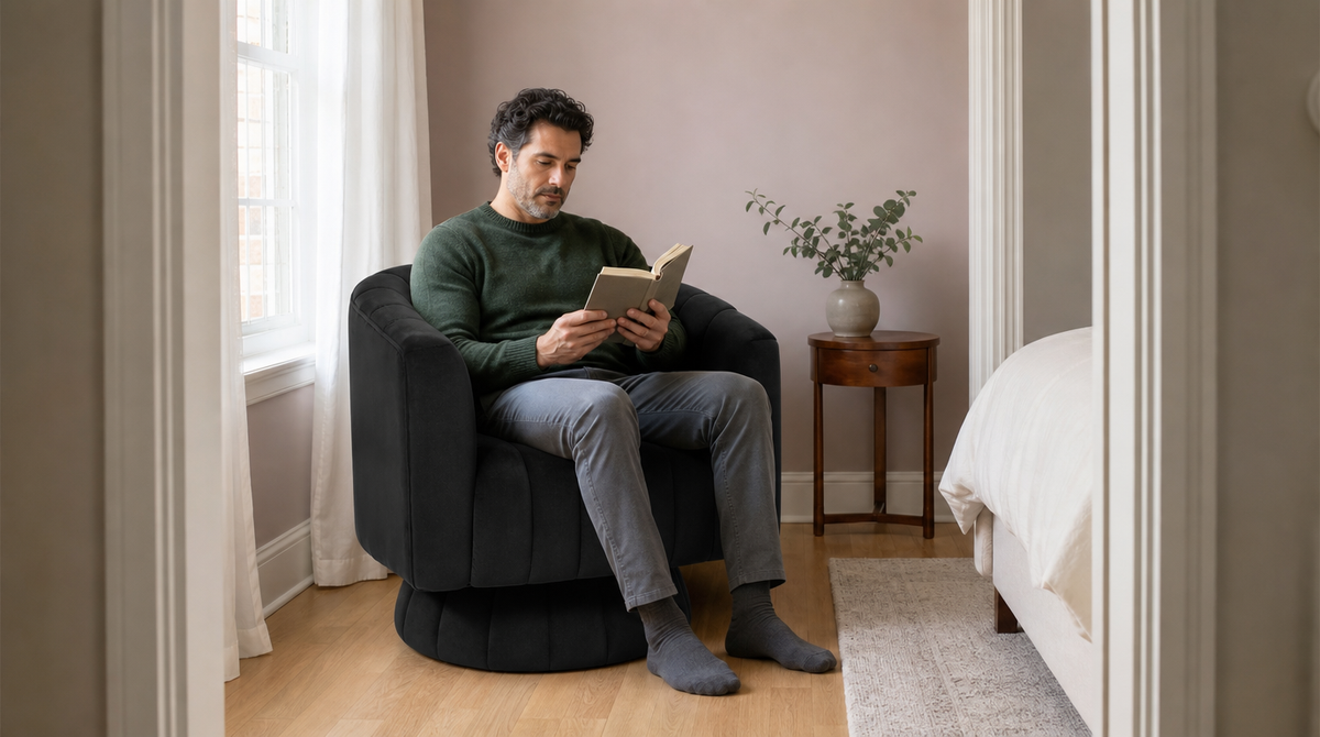 “Man reading in a black barrel chair beside a bedroom window, with both feet resting on the floor.”
