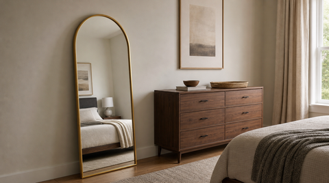 “Gold arched floor mirror leaning against a bedroom wall beside a wide wooden dresser, reflecting the bed.”
