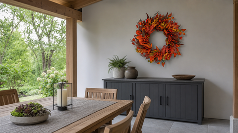 Red-orange leaf and berry wreath above a dark cabinet beside a wooden dining table in a covered outdoor space.