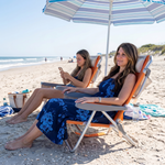 Two people relaxing in matching light blue stripe backpack beach chairs on the sand, each reclined with a drink in hand and the ocean in the background