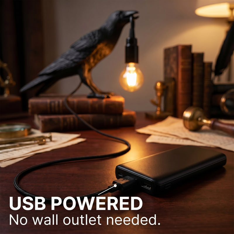 Black raven lamp on a wood desk connected to a USB power bank among vintage books and a magnifying glass