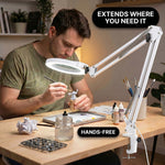 Man using a clamp-mounted LED magnifying lamp to hand-paint a miniature figure at a hobby desk