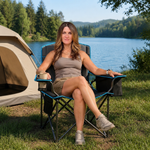 Woman relaxing in a black oversized camping chair by a lake with a water bottle in the cup holder beside a tent