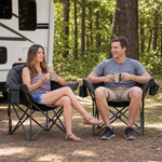 A man and woman relaxing in a matching pair of oversized camping chairs at a wooded campsite with tents and a campfire