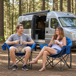 A man and woman relaxing in a matching pair of oversized camping chairs at a wooded campsite with tents and a campfire