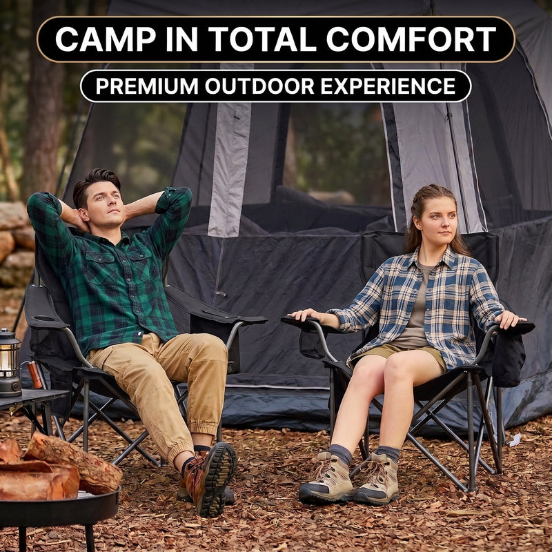 A man and woman relaxing in a matching pair of oversized camping chairs at a wooded campsite with tents and a campfire