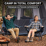 A man and woman relaxing in a matching pair of oversized camping chairs at a wooded campsite with tents and a campfire