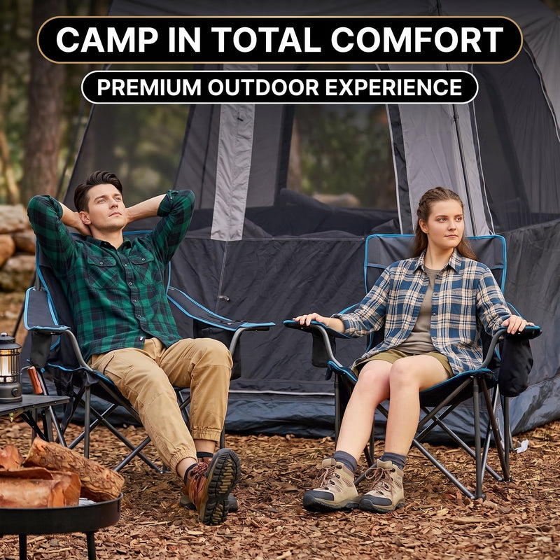 A man and woman relaxing in a matching pair of oversized camping chairs at a wooded campsite with tents and a campfire