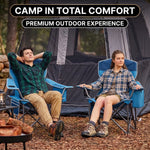 A man and woman relaxing in a matching pair of oversized camping chairs at a wooded campsite with tents and a campfire