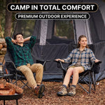 A man and woman relaxing in a matching pair of oversized camping chairs at a wooded campsite with tents and a campfire
