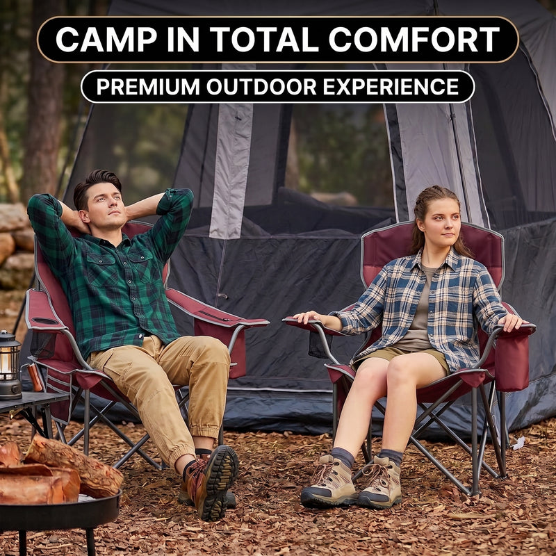 A man and woman relaxing in a matching pair of oversized camping chairs at a wooded campsite with tents and a campfire