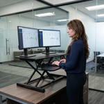 Woman standing at a black standing desk converter with dual monitors in a modern office