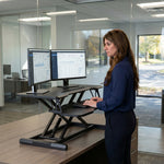 Woman standing at a black standing desk converter with dual monitors in a modern office