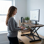 Woman standing at a black standing desk converter with dual monitors in a modern office
